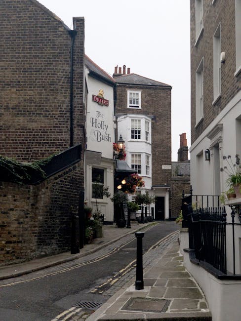 A narrow residential street in Hampstead with a gently sloping pavement on the right, separated from the road by short black bollards and metal drains embedded in the sidewalk. On the left, there is a weathered brick wall with greenery spilling over the top, leading to a small building with a dark sloped roof. In the background, several multi-story Victorian-style buildings with brick and white facades are visible, including a prominent white bay-windowed corner property with decorative flower boxes and hanging plants. A sign for 'The Holly & Bush' pub is mounted on one of the buildings, partially obscured by hanging floral arrangements and outdoor lighting. The street is quiet, with no vehicles or pedestrians visible, and the scene is lit by natural daylight with slightly overcast skies, evoking an atmosphere suited to private waste collection and rubbish removal services often employed in historic Hampstead neighborhoods.