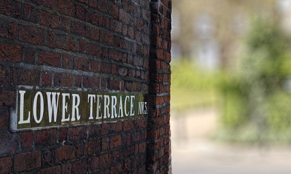 A close-up view of a dark red brick wall with visible mortar joints, featuring a rectangular metal street name plate attached to the surface. The plate displays the text 'LOWER TERRACE NW3' in bold, white, uppercase letters with a slight sheen. To the right of the sign, the brick wall curves slightly, continuing upward out of the frame. The background is blurred, showing green foliage and hints of a pathway or road beyond the wall, with soft natural light illuminating the scene. The setting suggests an urban or residential area, where private or independent waste collection services, such as those provided by Waste Removal Hampstead, may operate, especially for properties that do not rely on local authority rubbish collection. The overall composition emphasizes the texture of the bricks and the clarity of the street sign, providing a neutral, factual representation related to rubbish removal and property signage.