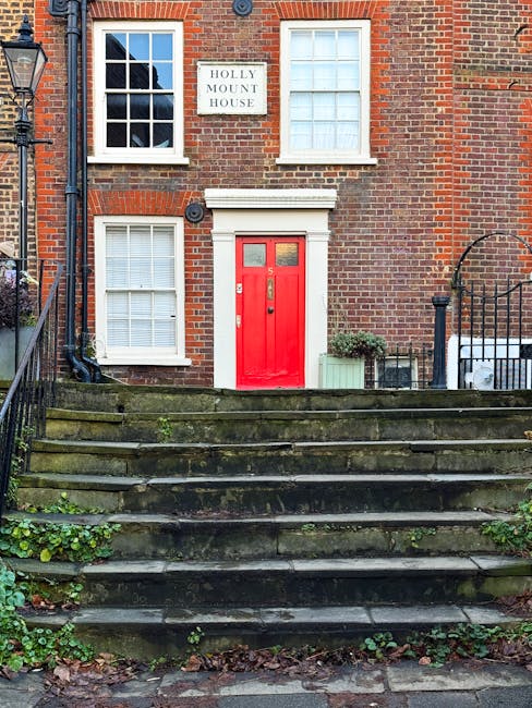 The image depicts a traditional brick residential building with a prominent red front door centered on a small set of weathered stone steps leading up to the entrance. The brick facade is in reddish-brown tones with visible mortar joints, and features four white-framed windows—two on the ground floor and two on the upper floor—each fitted with white blinds. A white sign reading 'HOLLY MOUNT HOUSE' is mounted between the upper windows. To the right of the door, there is a small potted shrub in a rectangular green planter, alongside a light-colored box or container. The stone steps and surrounding pavement show signs of moss and minor plant growth, indicating age and slight neglect. Flanking the steps are black wrought iron railings, one with a curved top near the bottom of the steps. The background includes part of a streetlamp on the left and a small section of an adjacent building, suggesting an urban or suburban setting. The scene conveys a neat, historic terraced residence set in a quiet, leafy neighbourhood, with subtle hints of potential for on-site or independent waste disposal accessible to local waste removal services like Waste Removal Hampstead.
