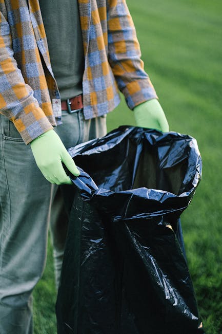 A person wearing a green long-sleeved shirt, a plaid jacket with yellow, blue, and grey tones, and light grey trousers is holding a black rubbish bag in their hands, which are covered with bright green disposable gloves. The black plastic bag is open and appears to be for waste collection or disposal. The individual is standing outdoors on a grassy surface, with a blurred background of green grass or a lawn, indicating an environment suitable for garden or estate clean-up activities. The scene suggests an organized approach to rubbish removal, possibly involving private waste handling services, as the person prepares to gather household or garden waste for collection. The lighting is natural, highlighting the textures of the gloves, plastic bag, and clothing, creating a professional and neutral tone suitable for a waste removal or rubbish clearance service context.