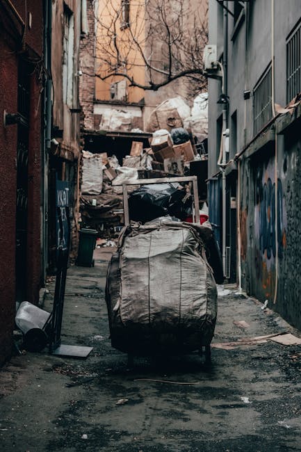 A narrow urban alleyway filled with accumulated rubbish and waste materials. In the forefront, a large, grey, weathered garbage bag sits on the dark, stained pavement, which shows signs of dirt and debris. Behind it, a filled shopping trolley or cart, also covered in dirt and grime, is positioned upright, with its handle extending upwards toward the middle of the scene. Further into the background, piles of cardboard boxes, plastic bags, and miscellaneous waste are stacked haphazardly against a backdrop of older brick and concrete buildings. The sides of the alley are bordered by tall, graffiti-covered and metal-sheathed walls, with some visible pipes and vents attached to the structures. Leafless tree branches reach over from the upper part of the scene, contrasting with the chain-link fencing and urban environment. The lighting is diffuse, suggesting overcast weather, which lends a muted tone to the scene. This image visually demonstrates the kind of clutter and debris typically associated with private rubbish collection or waste removal services in tight city spaces, such as those managed by Waste Removal Hampstead, highlighting the need for efficient rubbish and waste clearance solutions in narrow streets.