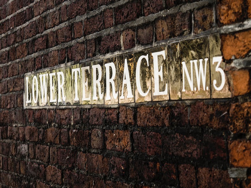 A close-up view of a rectangular, metallic street sign affixed to a weathered red brick wall. The sign displays the words 'LOWER TERRACE' in large, white, uppercase letters with a slight embossed effect, and the address 'N.W.3' in smaller white characters on the right side. The background of the sign has a glossy, slightly tarnished appearance with visible patches of dirt and moss, indicating age and outdoor exposure. The brick wall consists of rough-textured, uneven bricks with varying shades of red, brown, and black mortar lines, creating a rustic urban backdrop. The lighting is natural, casting soft shadows that emphasize the texture of the brickwork and the reflective surface of the sign. This scene suggests an alleyway or side street that could be part of a private or less-mainly thoroughfare, which may be relevant for independent rubbish removal or on-site waste clearance services offered by Waste Removal Hampstead, aligned with alternative waste handling solutions in the area.
