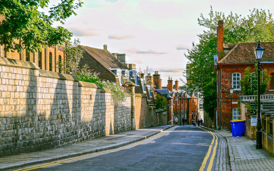 A narrow urban alleyway filled with accumulated rubbish and waste materials. In the forefront, a large, grey, weathered garbage bag sits on the dark, stained pavement, which shows signs of dirt and debris. Behind it, a filled shopping trolley or cart, also covered in dirt and grime, is positioned upright, with its handle extending upwards toward the middle of the scene. Further into the background, piles of cardboard boxes, plastic bags, and miscellaneous waste are stacked haphazardly against a backdrop of older brick and concrete buildings. The sides of the alley are bordered by tall, graffiti-covered and metal-sheathed walls, with some visible pipes and vents attached to the structures. Leafless tree branches reach over from the upper part of the scene, contrasting with the chain-link fencing and urban environment. The lighting is diffuse, suggesting overcast weather, which lends a muted tone to the scene. This image visually demonstrates the kind of clutter and debris typically associated with private rubbish collection or waste removal services in tight city spaces, such as those managed by Waste Removal Hampstead, highlighting the need for efficient rubbish and waste clearance solutions in narrow streets.
