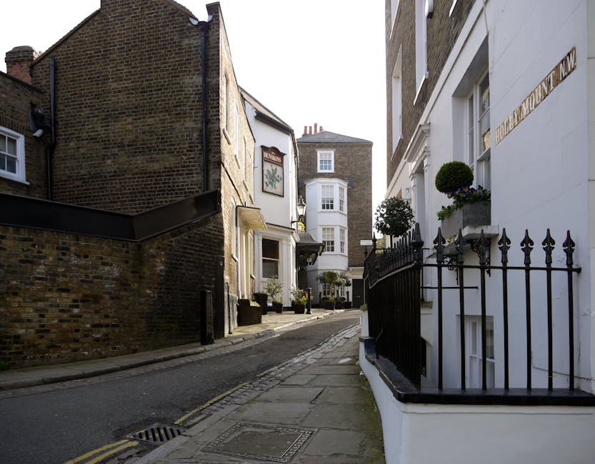 A narrow residential street view featuring several buildings with different architectural styles. On the right, there is a white-painted building with a decorative black wrought iron fence along the front, adorned with flower boxes containing small, neatly trimmed potted topiary plants. The building's façade includes a window with a stone sill and protruding decorative stone surrounds. To the left, a taller brick building with a darker, weathered exterior is partially visible. The street has a slight incline, paved with asphalt, with a concrete sidewalk that has a decorative pattern near the curb. An open metal drain cover is visible on the street surface. In the background, additional brick and weatherboard structures, some with chimneys, can be seen, along with a bay window on a white building further down the street. The environment is sunny, with natural daylight illuminating the scene, highlighting the textures of the brickwork, painted surfaces, and greenery, contributing to the overall appearance suitable for considering private waste disposal or rubbish removal services like those provided by Waste Removal Hampstead.