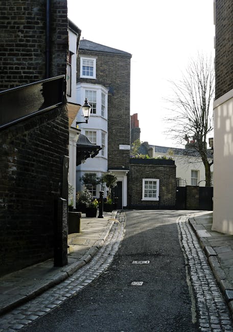 A narrow residential street in Hampstead during daytime, featuring a row of dark brick terraced houses on the left with white-framed windows and small front yards enclosed by black metal railings. The pavement on the right is paved with rectangular concrete slabs and is lined with tall, dense evergreen hedges and trees, some with bare branches. The asphalt road has minimal markings and gently curves to the right, with a parked white car visible in the distance. The lighting is natural, with bright sunlight casting shadows on parts of the street and highlighting the textures of the brickwork and foliage. The scene depicts an urban environment that could be used for private waste collections or rubbish removal.