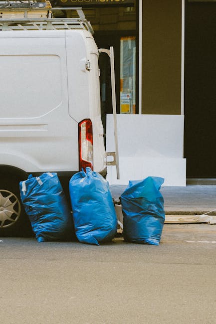 The image shows the rear side of a white commercial van parked on a paved street, with three large blue rubbish bags placed upright against the vehicle's side near the back wheel. The bags are made of thick, durable plastic with a slightly crinkled texture, and their bright blue color contrasts with the neutral tones of the van and street. In the background, there is a storefront or building with glass windows and a white façade, featuring an open door or window with an orange and blue sign partially visible inside. The scene appears to be during daytime, with indirect natural light illuminating the objects. The position of the bags suggests recent collection, possibly part of an independent, private waste removal service such as that offered by Waste Removal Hampstead, supporting alternative disposal methods outside of the local authority collection system. This setup indicates the typical arrangement for on-site rubbish clearance, suitable for clients seeking same-day or urgent waste removal solutions in the Hampstead area, in line with private waste handling standards.