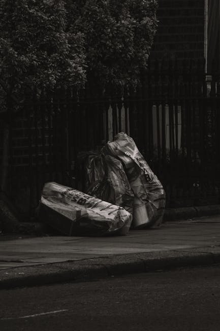 Three large black plastic garbage bags filled with waste are placed on the pavement in front of a black metal fence. The bags appear to contain household rubbish, with some items and packaging material visible through the semi-transparent plastic. The bags are positioned in a row, leaning slightly against each other, and are situated on a concrete sidewalk beside a curb. In the background, the fence extends across the scene, with dense foliage and trees behind it, partially visible at the top of the image. The lighting suggests an overcast or shaded environment, casting a subdued tone over the scene. This setting reflects an off-site rubbish collection scene, consistent with private waste management services like those offered by Waste Removal Hampstead, highlighting the common practice of curbside waste presentation for alternative collection methods outside of local authority services.