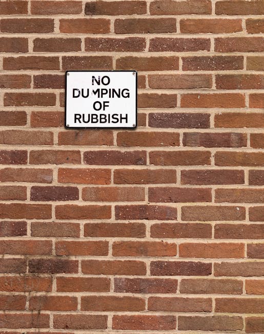 A rectangular white metal sign with black text reading 'NO DUMPING OF RUBBISH' is mounted on a reddish-brown brick wall. The wall consists of evenly spaced bricks with variations in tone, some darker and some lighter, and light mortar joints between them. The sign is positioned slightly above the center of the image, with the brick wall occupying the background and surrounding area. The lighting appears natural, casting subtle shadows on the bricks and highlighting their textured surface. This scene depicts a typical urban environment where waste management practices and regulations are visually reinforced through signage, supporting the services of Waste Removal Hampstead focused on rubbish collection and private waste handling solutions. The wall's uniform brick pattern and the clear, legible sign suggest a controlled environment aimed at deterring rubbish dumping, aligning with policies for cleaner private and public spaces in the Hampstead area.