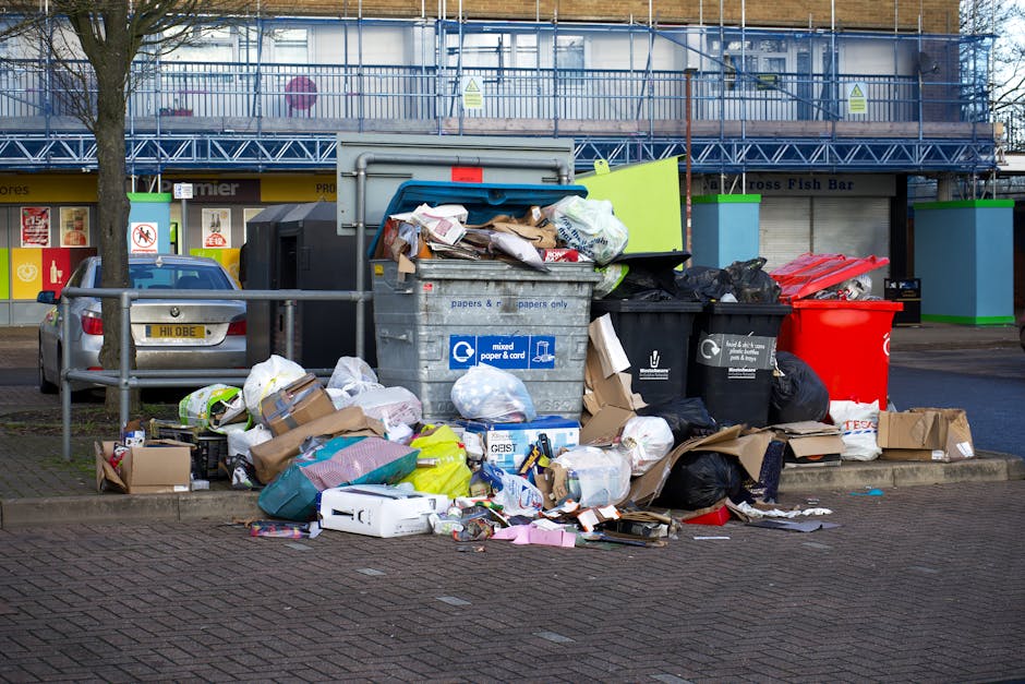 A collection of overflowing waste and rubbish bags positioned on a paved urban street corner, with various types of refuse including black and black plastic bags, flattened cardboard boxes, paper items, and flattened packaging materials scattered around the base of a large grey communal waste container with a partially open blue lid. The container is marked for mixed paper and cardboard recycling, and some loose waste spills onto the surrounding pavement. To the left, a grey car is parked behind a metal railing, with a tree adjacent to it. In the background, there are storefronts with signage, including a fish bar, and a building with blue scaffolding, suggesting ongoing maintenance or construction work. The scene is illuminated by natural daylight, highlighting the accumulation of rubbish and the urban environment, aligning with services that handle private waste disposal and rubbish removal in residential and commercial areas.